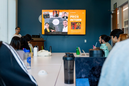 A student gives a presentation about an internship to a group seated at a long table. A large screen displays "PROX INTERN" and various photos.
