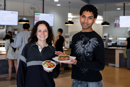 wo smiling students hold up their lunch—a salad bowl and a sandwich—while standing in a modern university cafeteria with digital menu boards.