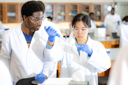 A male and female student wearing white lab coats and blue gloves perform a science experiment in a lab.
