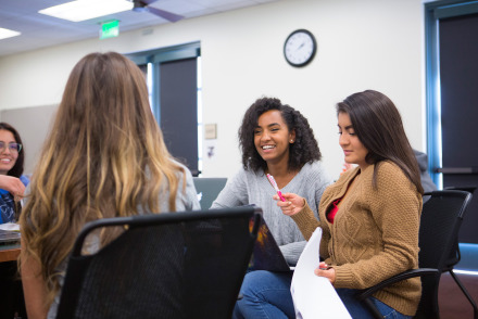 A group of female students sits around a table in a brightly lit classroom, engaged in a discussion. One student with long blonde hair faces away from the camera, while two other students opposite her listen and smile, with one holding a pink pen and papers.