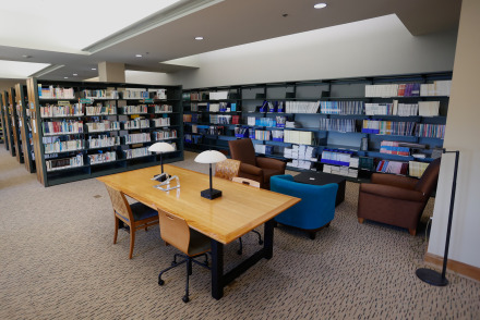 Bookshelves surround a wooden desk with two chairs as well as an assortment of blue and brown sofa chairs inside the library.