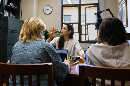 A female professor gestures while mentoring two students at a desk with laptops and fruit.