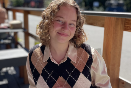 A woman with curly dirty blonde hair wears a checkered vest and smiles while sitting on a patio outside with a blue sky and red umbrella above.