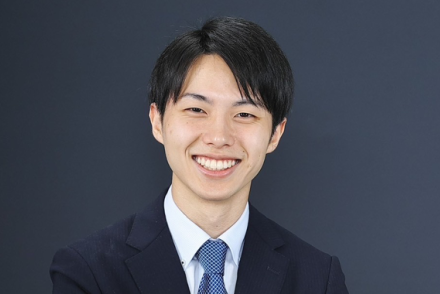 A man with dark brown hair wearing a dark suit with a lighter blue tie smiles.