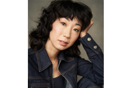 A woman with curly dark brown hair smiles and tilts her head to the right. She is wearing a jean jacket and resting her head in her left hand.