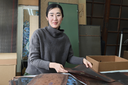 A woman with black, pulled-back hair, wears a grey turtleneck sweater and is working with a pile of Japanese textiles at a table. 
