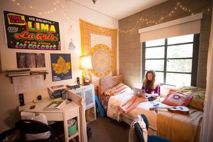 A student sits on a bed using a laptop in a cozy dorm room decorated with string lights, colorful posters, a tapestry, and a ukulele.