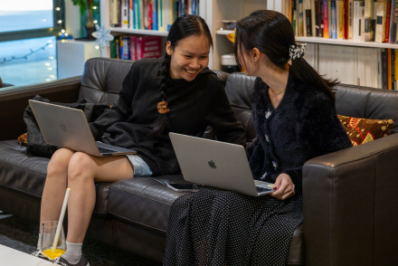 Two students with long black hair sit on a couch with their laptops on their laps and share a laugh.
