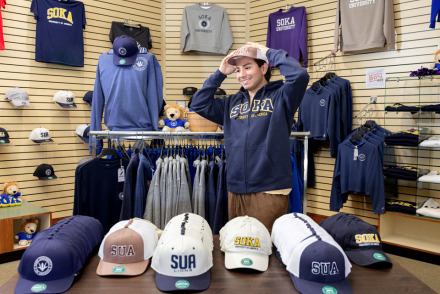 A student tries on an SUA hat and Soka sweatshirt in the bookstore surrounded by other merchandise including hats and attire.