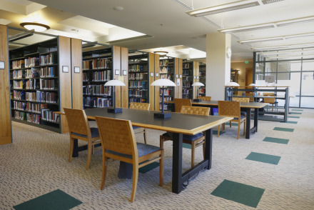 Tables with two desk lamps and four wooden chairs are aligned to the right of rows of floor to ceiling bookshelves inside the library.