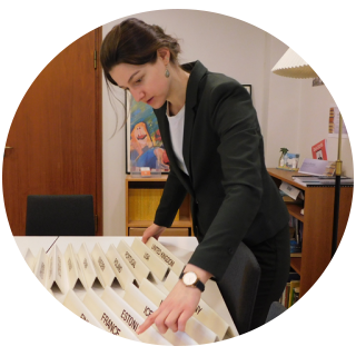 MacKenzie Kermoade is wearing a suit jacket and thumbing through various countries' nameplates in a meeting room.