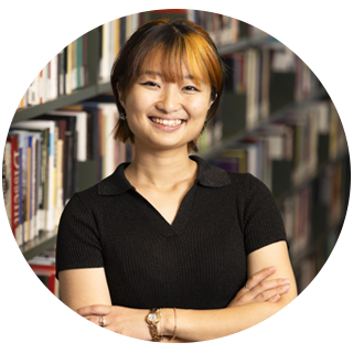 A woman with orange and black hair, wearing a black v-neck stands with her arms folded and smiles in front of bookshelves in the library.
